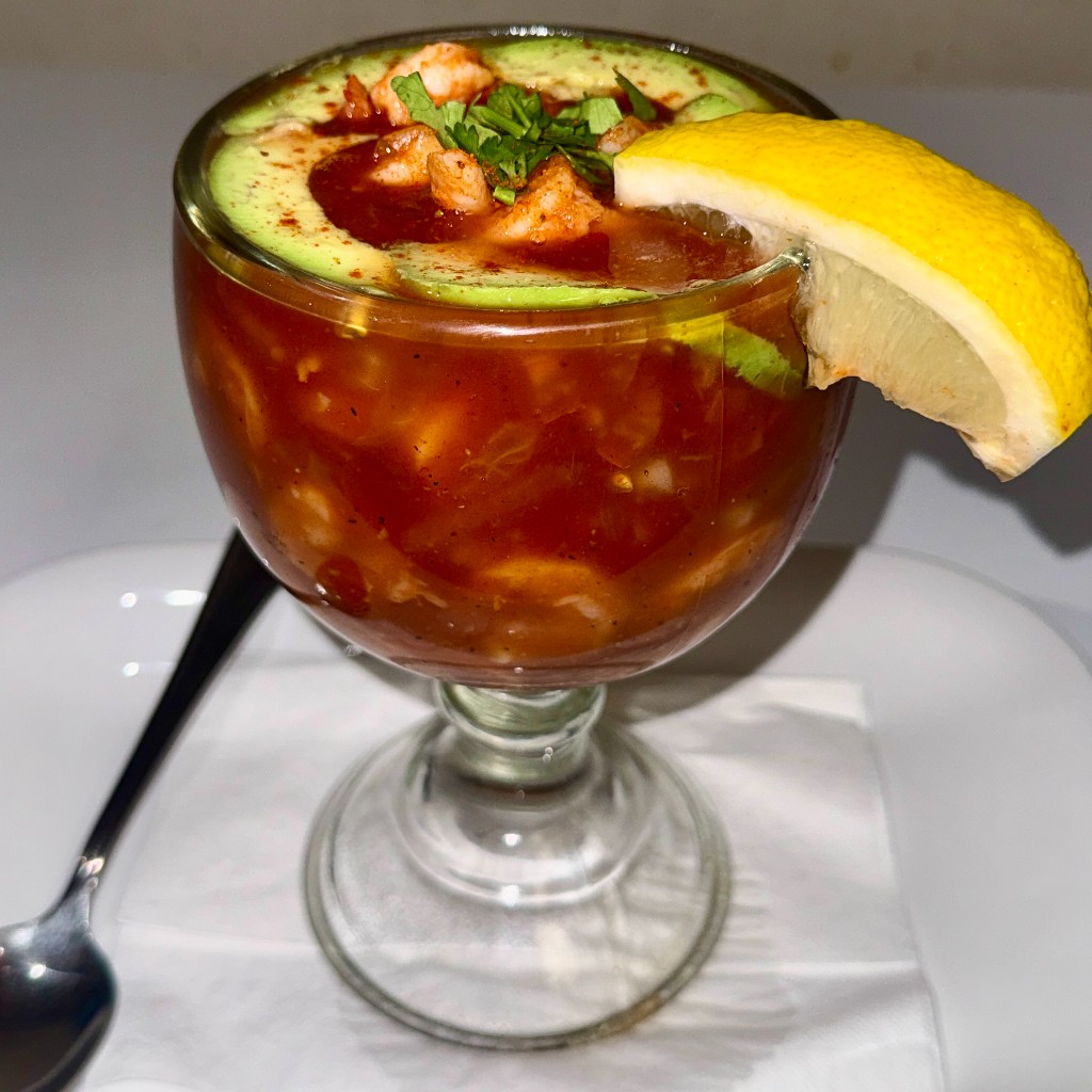 A shrimp cocktail in a glass, topped with avocado, herbs, and a lemon wedge
