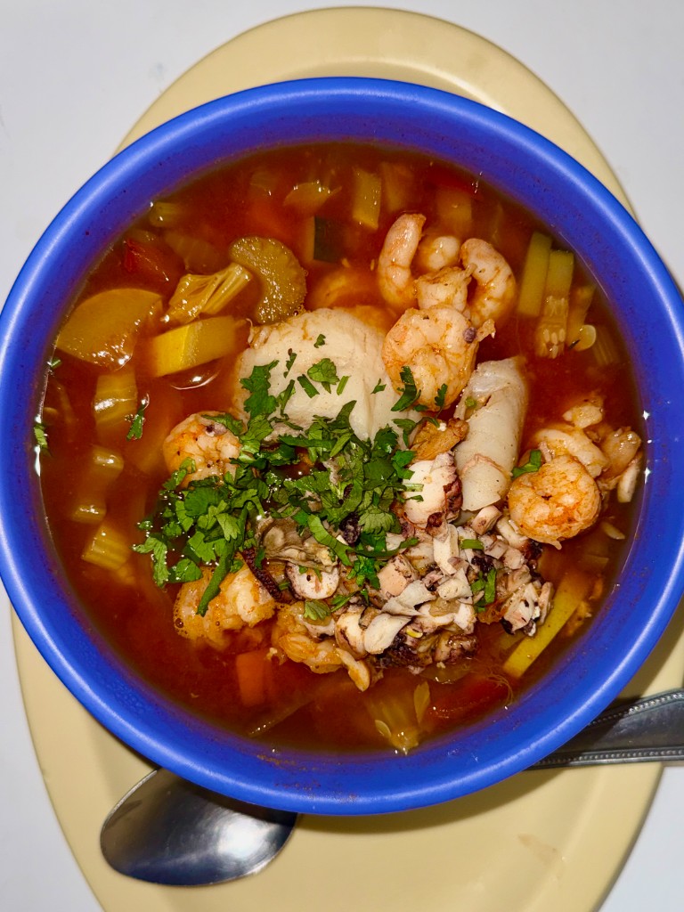 A blue bowl of seafood soup with shrimp, herbs, and vegetables